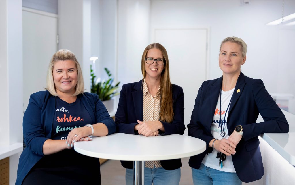 Kolme naista seisomassa pöydän takana, kasvot kameraan päin.